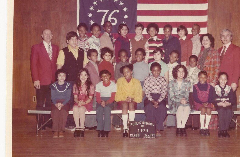 A vintage class photo from 1976 shows a diverse group of students and teachers, embodying early principles of social emotional learning. They pose proudly in the school auditorium before a banner with "76" and stars, alongside a U.S. flag, representing Public School 116, Class 5-8 IS.