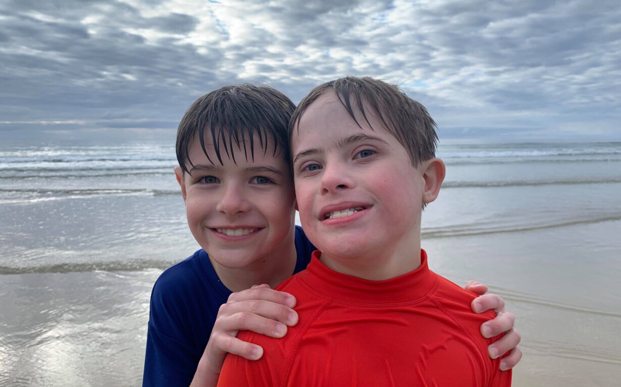 Two smiling children at the beach stand close together in the shallow water, embodying social emotional learning through shared joy. One wears a blue shirt and the other a red shirt. Overcast skies and gentle waves are in the background, with a bird flying above.