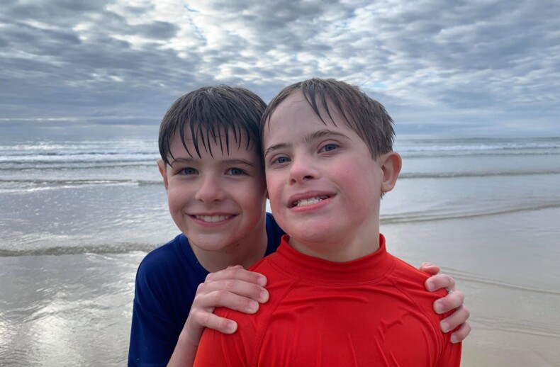 Two smiling children at the beach stand close together in the shallow water, embodying social emotional learning through shared joy. One wears a blue shirt and the other a red shirt. Overcast skies and gentle waves are in the background, with a bird flying above.