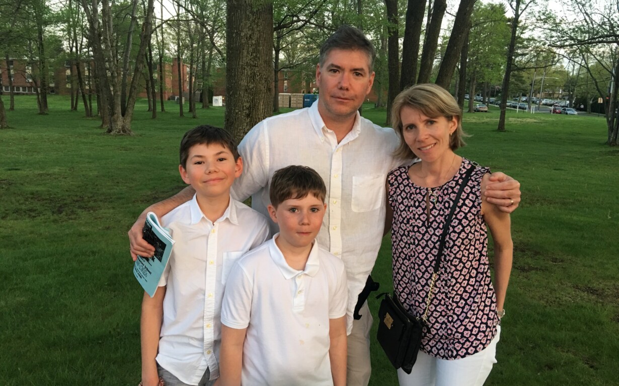 A family of four stands together in a park. They are wearing casual clothes, with the two children in front and the parents behind them, enjoying an afternoon filled with SEL activities. Trees and grass fill the background, creating a serene outdoor setting.