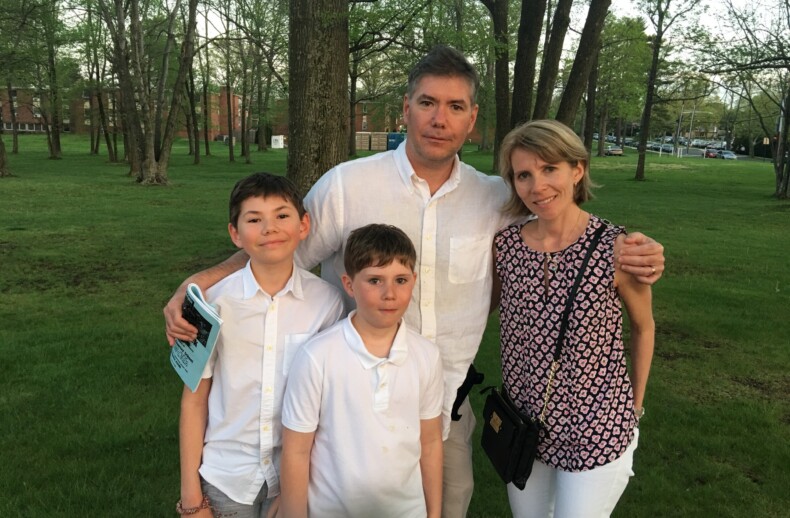 A family of four stands together in a park. They are wearing casual clothes, with the two children in front and the parents behind them, enjoying an afternoon filled with SEL activities. Trees and grass fill the background, creating a serene outdoor setting.