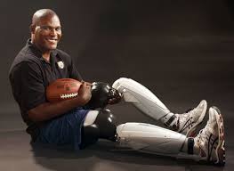 A man with prosthetic legs and one prosthetic arm is sitting on the floor, holding a football. He is smiling, wearing a black shirt, dark shorts, and sneakers against a neutral dark background—an inspiring image perfect for SEL activities that promote resilience and positivity.