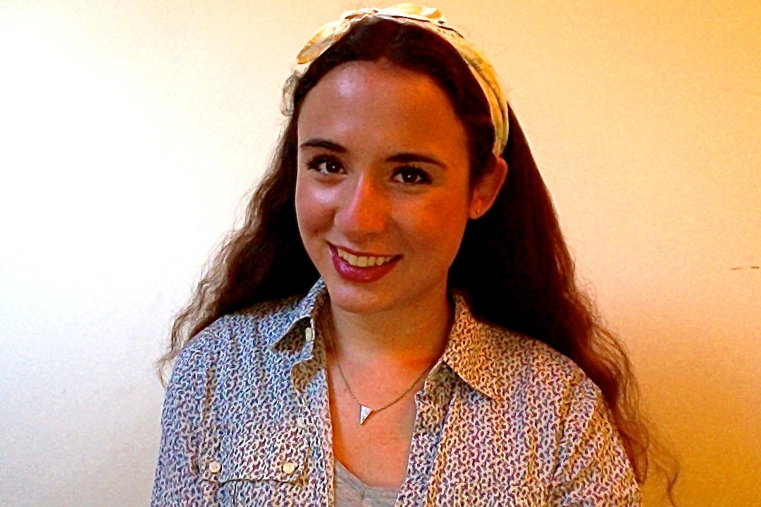 A woman with long, wavy hair smiles at the camera, embodying the positivity of social emotional learning activities. She is wearing a patterned shirt, a necklace, and a headband. The background is a plain, light-colored wall.