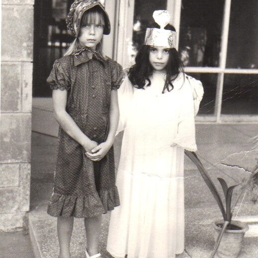 Two young girls, embodying themes from a social emotional learning curriculum, stand side by side in front of a building. The girl on the left wears a patterned dress and headscarf, while the one on the right sports a crown and light-colored gown. Both have serious expressions in this black-and-white photo.