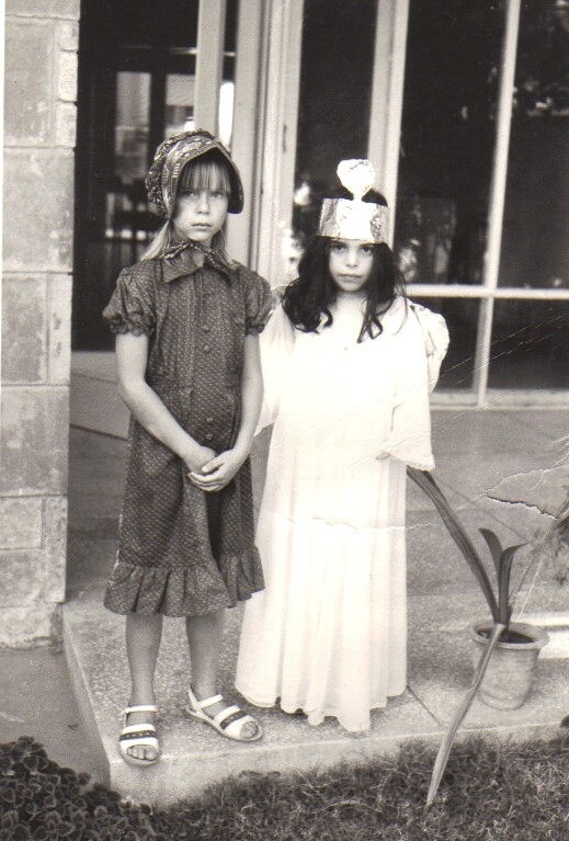 Two young girls, embodying themes from a social emotional learning curriculum, stand side by side in front of a building. The girl on the left wears a patterned dress and headscarf, while the one on the right sports a crown and light-colored gown. Both have serious expressions in this black-and-white photo.