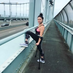 A woman with a prosthetic leg stands confidently on a bridge, embodying the spirit of diversity programs. Dressed in athletic wear, she leans on the railing with crutches in hand, one foot elevated. Cars pass by in the background, highlighting her strength and determination.