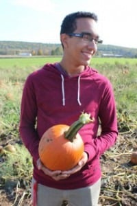A person in a maroon hoodie smiles while holding a small pumpkin, embodying the spirit of social emotional learning activities. They stand in a pumpkin patch with green fields and a forested hill in the background under a clear sky.