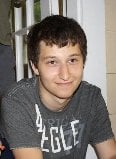 A person with short, dark hair is smiling. They are wearing a grey T-shirt with white lettering. The background features a window and indoor elements, creating a warm setting that encourages get to know you activities for students focused on building connections and understanding.