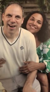 A smiling couple is seated together outdoors, radiating happiness and relaxation. The man wears a light sweater and shorts, while the woman dons a green and white patterned dress. Their warm embrace mirrors the harmony found in SEL activities, fostering emotional connections and well-being.