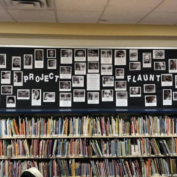 A library with a display titled "Project Flaunt" features a collage of black and white close-up photos of eyes, ears, noses, and mouths. Below the display are shelves filled with colorful books that support social emotional learning activities.
