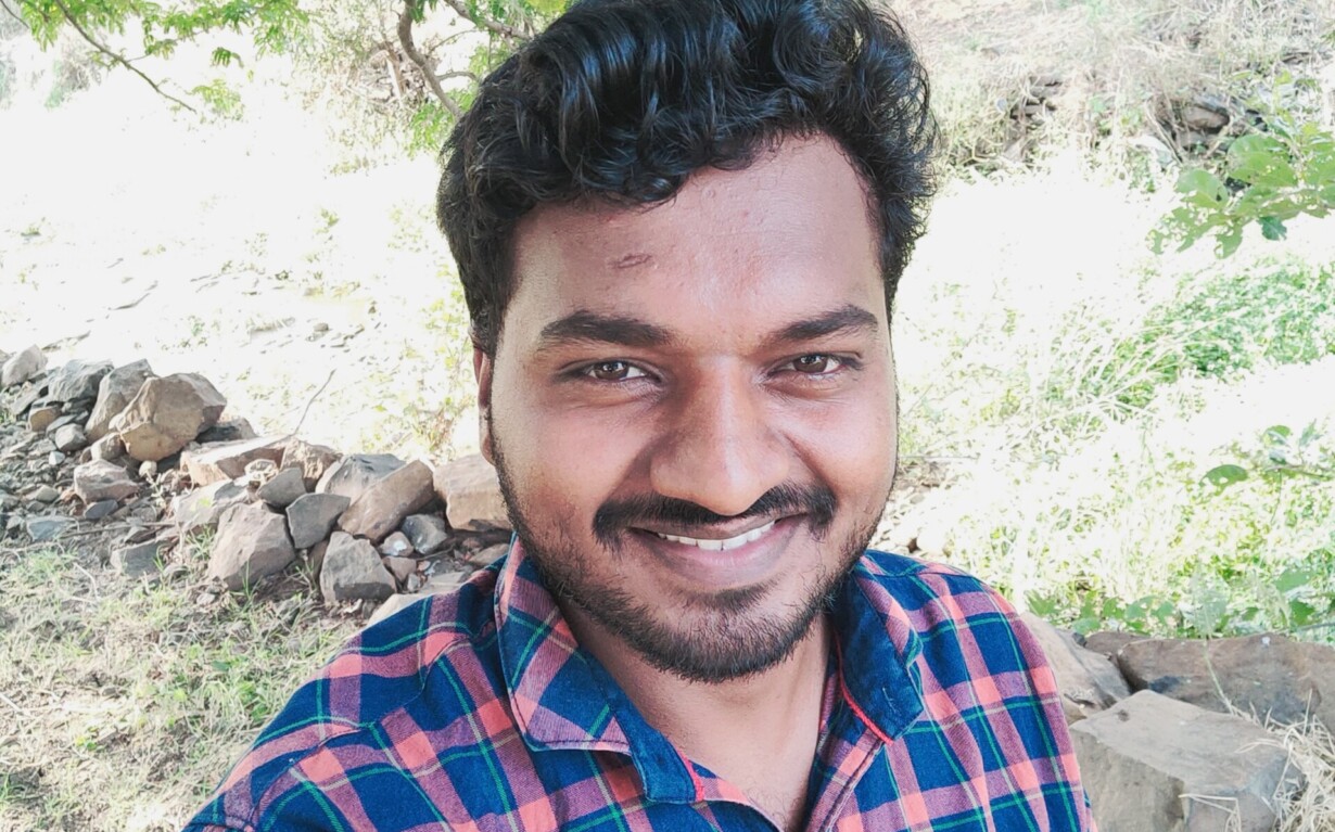 A man wearing a checkered shirt smiles at the camera while standing outdoors, embodying the positivity often found in SEL curriculum. He is surrounded by greenery and trees, with sunlight filtering through the leaves.