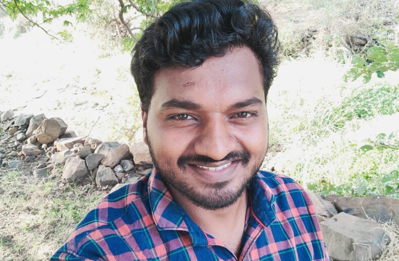 A man wearing a checkered shirt smiles at the camera while standing outdoors, embodying the positivity often found in SEL curriculum. He is surrounded by greenery and trees, with sunlight filtering through the leaves.