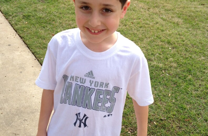 A young boy is standing outside on a sidewalk next to a grassy area, embracing the spirit of social emotional learning activities. He is smiling, wearing a white New York Yankees T-shirt and red shorts.
