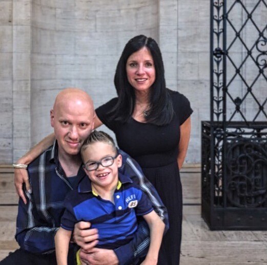 A family of three poses together indoors, embodying the spirit of SEL curriculum values. A man kneels, hugging a smiling young boy, while a woman stands behind with her hand on his shoulder. They're in front of a stone wall adorned with a decorative metal panel.