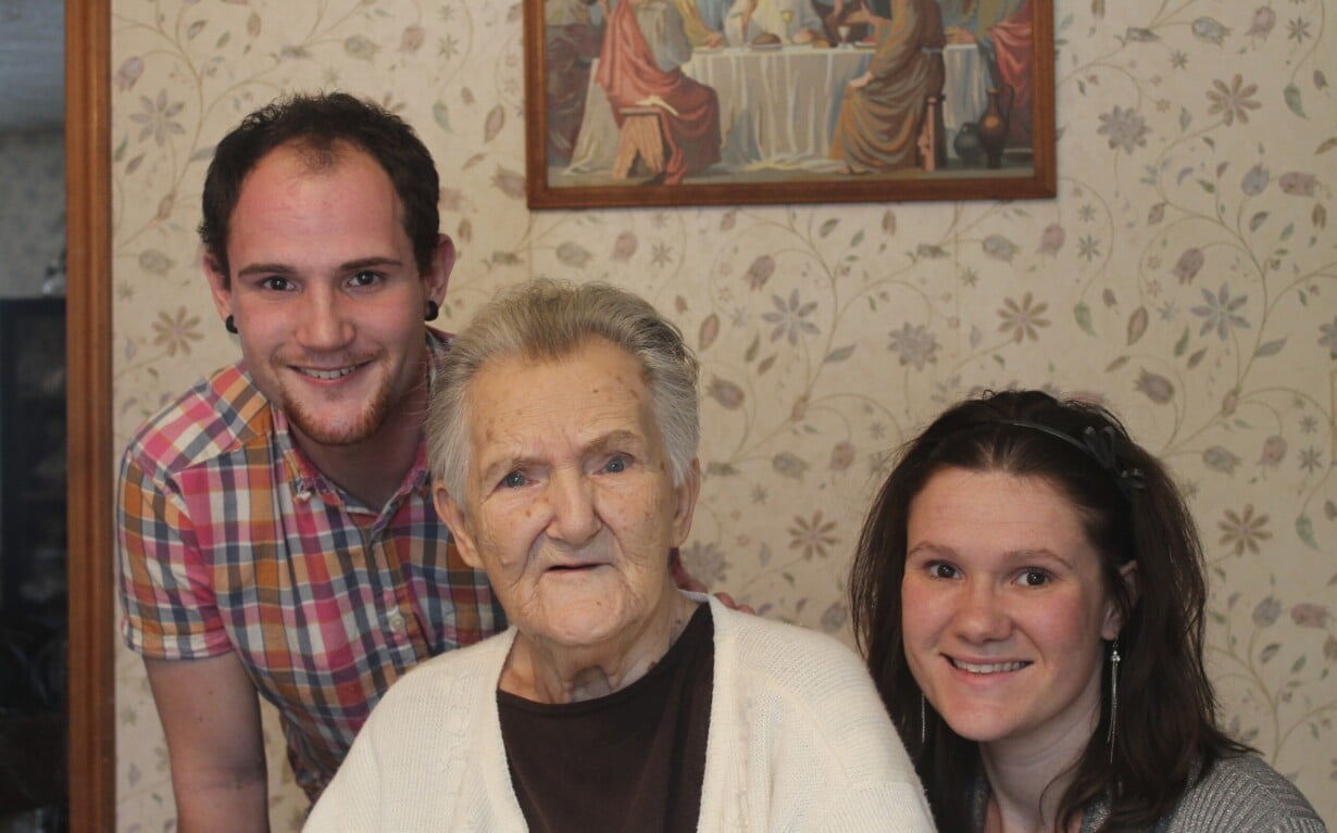 An elderly woman is seated between a young man and a young woman, all engaging in smiles that reflect their participation in social emotional learning activities. A painting hangs on the patterned wall behind them, adding to the harmonious scene.