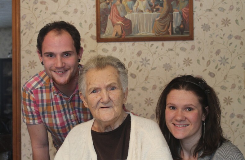 An elderly woman is seated between a young man and a young woman, all engaging in smiles that reflect their participation in social emotional learning activities. A painting hangs on the patterned wall behind them, adding to the harmonious scene.
