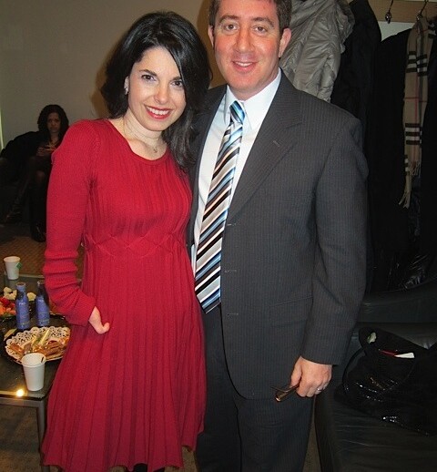 A woman in a red dress stands beside a man in a suit and tie, beaming smiles filling the cozy room. With coats on the wall and snacks nearby, it feels like one of those inviting get-to-know-you activities for students, fostering connections and warm conversations.