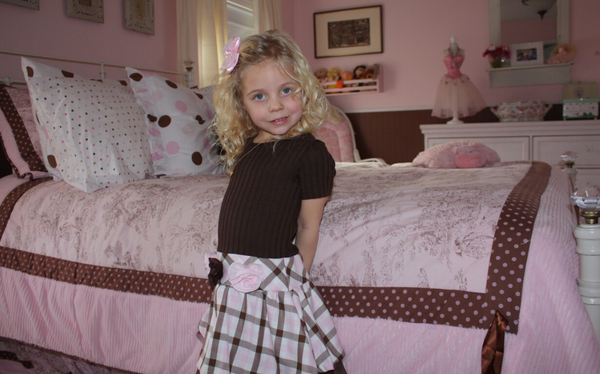 A young girl with blonde curly hair stands smiling in a pink and brown-themed bedroom. Wearing a brown top and plaid skirt with a pink bow, she embodies the joy found in SEL activities. The room features a pink bedspread with polka dots, a dresser, and soft toys filled with warmth.