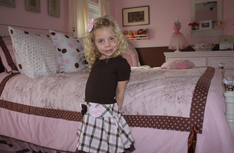 A young girl with blonde curly hair stands smiling in a pink and brown-themed bedroom. Wearing a brown top and plaid skirt with a pink bow, she embodies the joy found in SEL activities. The room features a pink bedspread with polka dots, a dresser, and soft toys filled with warmth.