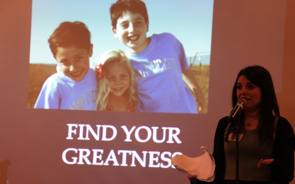 A woman speaks into a microphone in front of a projected image showing three smiling children outdoors, embodying the spirit of "FIND YOUR GREATNESS." This inspiring scene highlights the importance of social emotional learning activities in fostering connections.