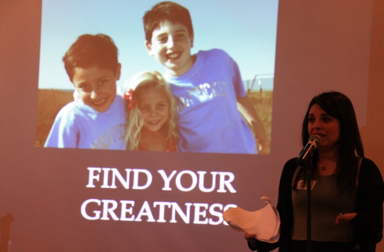 A woman speaks into a microphone in front of a projected image showing three smiling children outdoors, embodying the spirit of "FIND YOUR GREATNESS." This inspiring scene highlights the importance of social emotional learning activities in fostering connections.