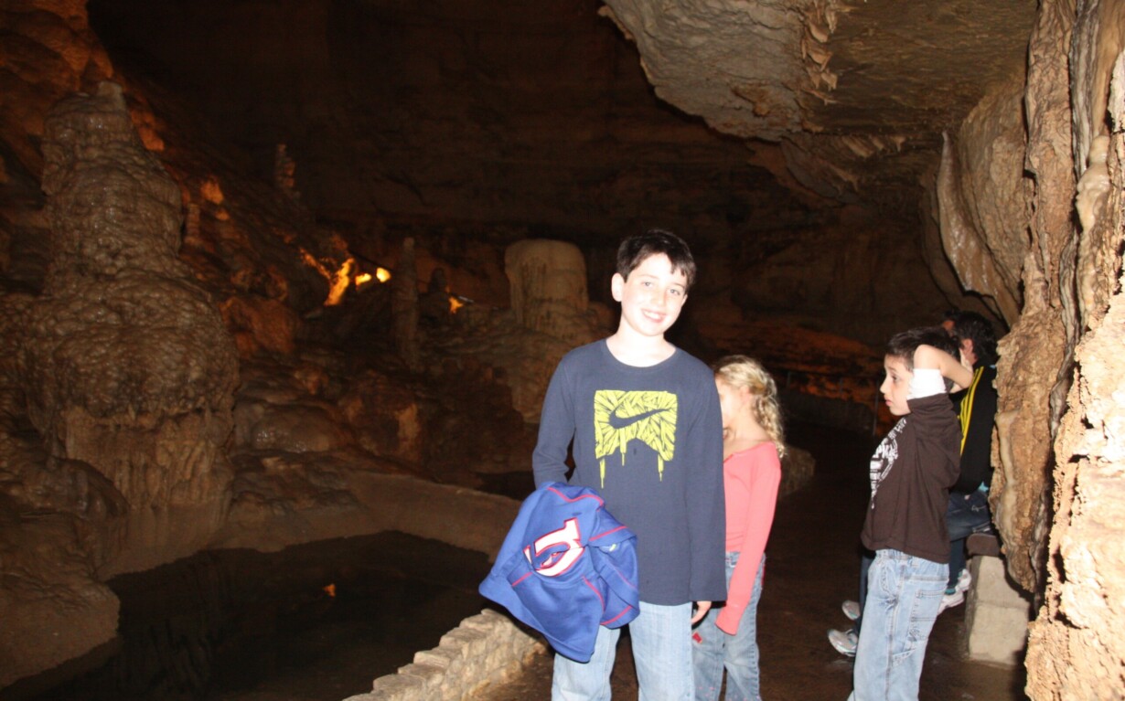 A group of children engaged in social emotional learning activities is exploring a dimly lit cave adorned with stalactites and stalagmites. One child in the foreground, smiling and holding a jacket, marvels at the rocky, uneven walls that frame a narrow walkway beside a small water body.
