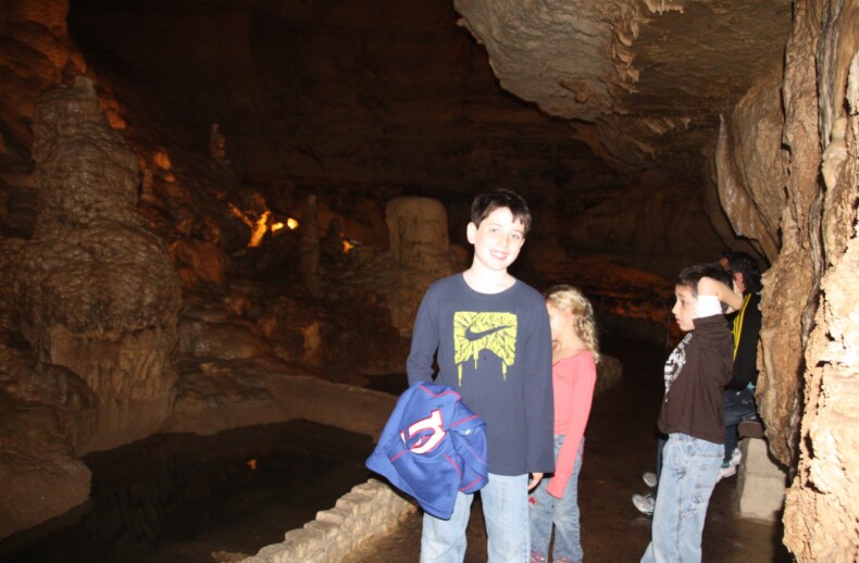 A group of children engaged in social emotional learning activities is exploring a dimly lit cave adorned with stalactites and stalagmites. One child in the foreground, smiling and holding a jacket, marvels at the rocky, uneven walls that frame a narrow walkway beside a small water body.