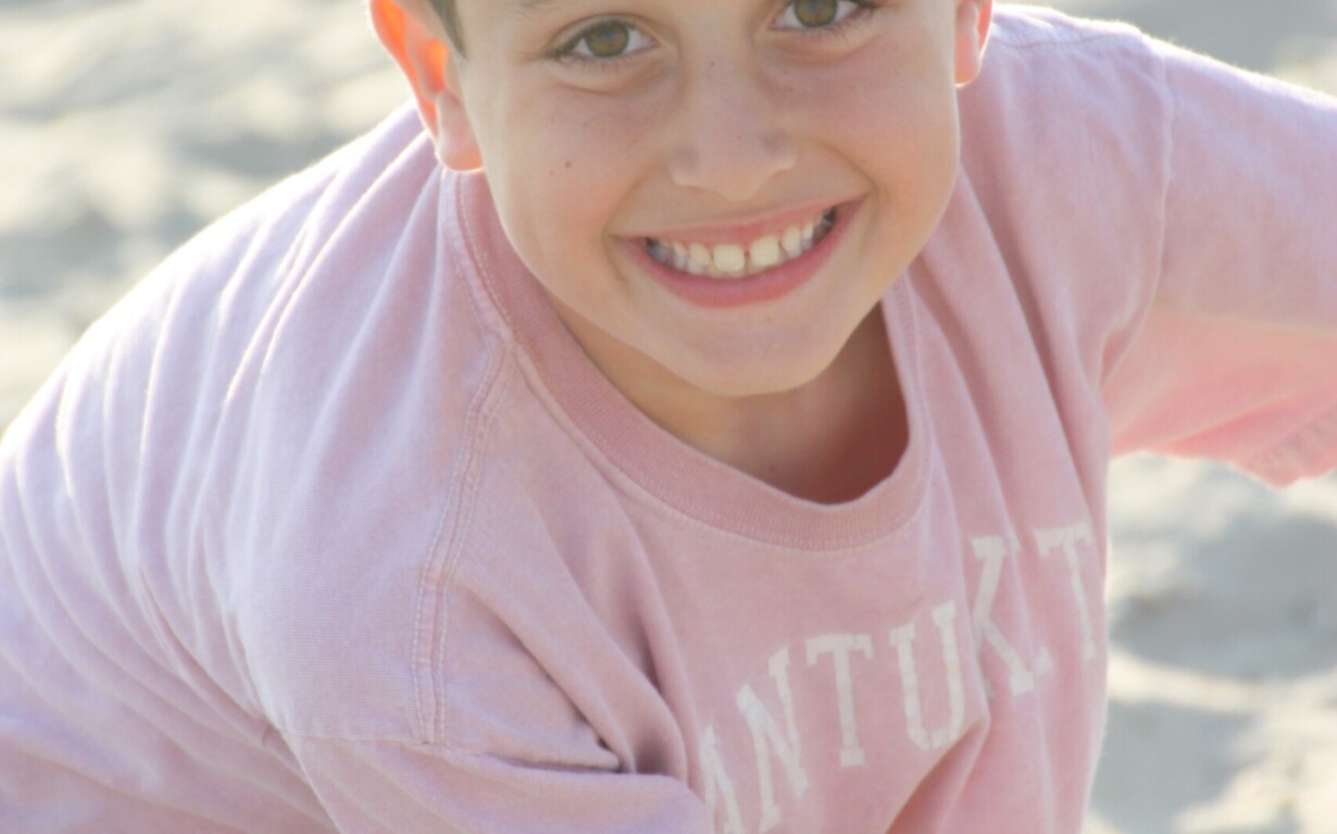 A young boy with short dark hair and a big smile is wearing a pink shirt. He is sitting on sandy ground, facing the camera with sunlight illuminating his face, embodying the joy often found in social emotional learning activities.