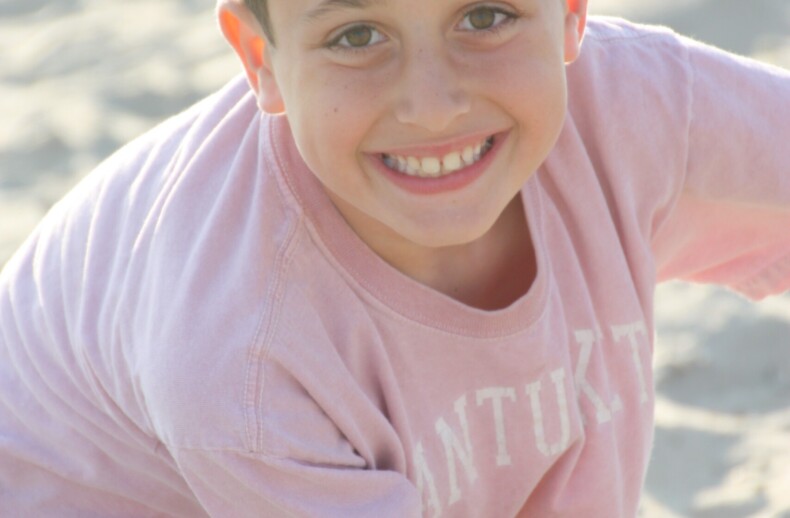 A young boy with short dark hair and a big smile is wearing a pink shirt. He is sitting on sandy ground, facing the camera with sunlight illuminating his face, embodying the joy often found in social emotional learning activities.