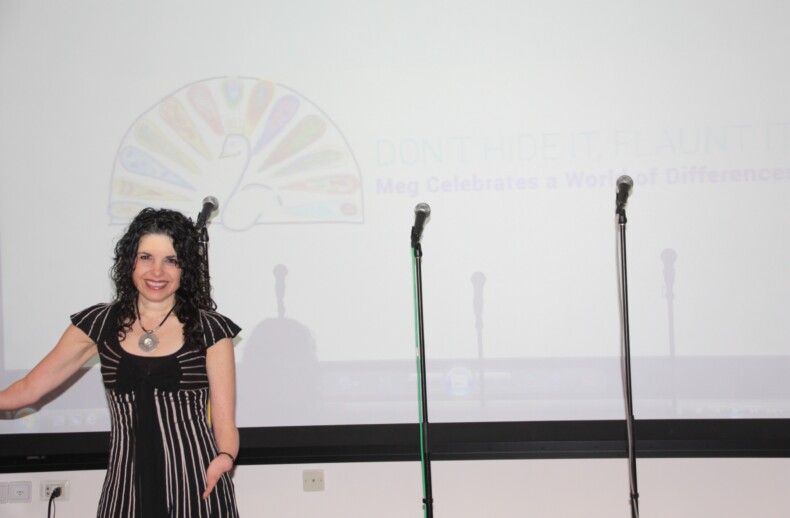 A woman with curly hair smiles while standing next to a white projection screen featuring a colorful peacock logo and partially visible text related to social-emotional learning activities. Two microphone stands are in front of the screen. She is wearing a striped black and white dress.