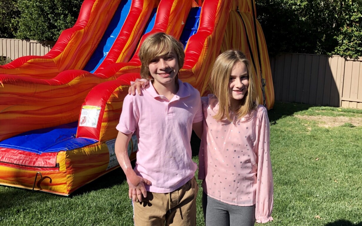 Two children stand smiling in front of a large inflatable slide, embodying the joy often cultivated in social-emotional learning activities. The boy on the left wears a light pink shirt and brown pants, while the girl on the right sports a pink sweater and gray pants. They're barefoot on a grassy yard with a wooden fence behind them.