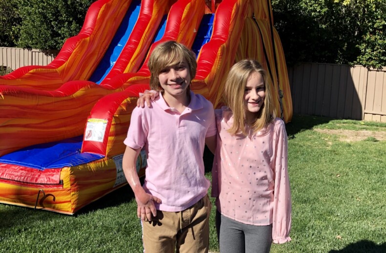 Two children stand smiling in front of a large inflatable slide, embodying the joy often cultivated in social-emotional learning activities. The boy on the left wears a light pink shirt and brown pants, while the girl on the right sports a pink sweater and gray pants. They're barefoot on a grassy yard with a wooden fence behind them.