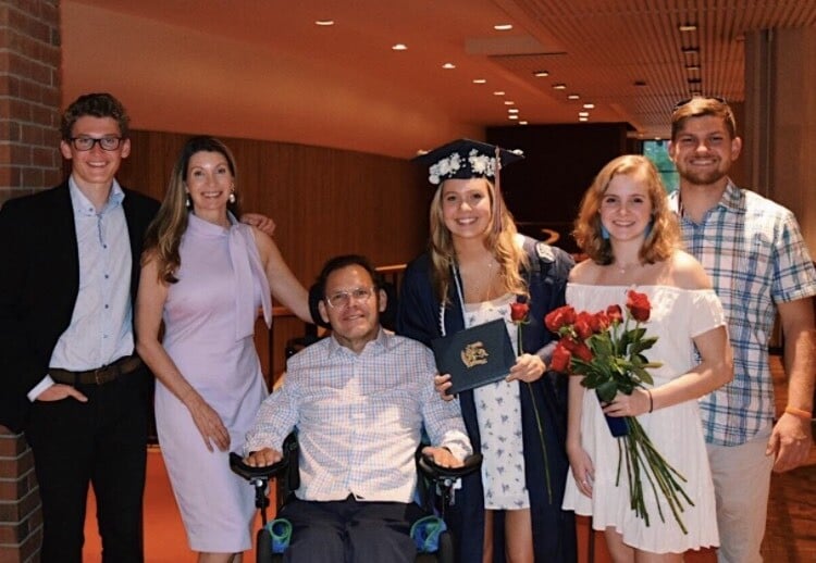 A group of six people posing indoors, celebrating achievements and reflecting the success of diversity programs. One person in the center is wearing a graduation cap and gown, holding a diploma and flowers. Another is in a wheelchair while others stand around them, all smiling warmly.