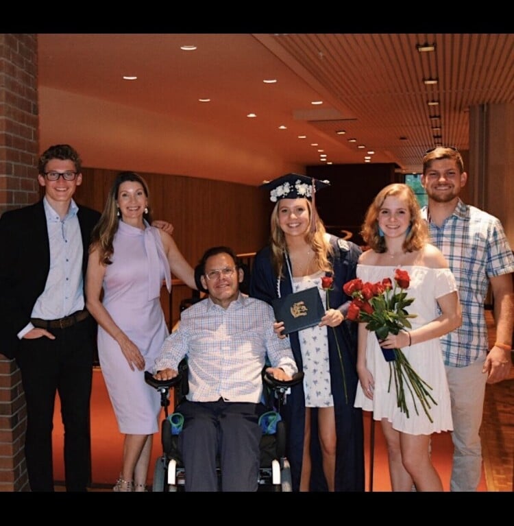 A group of six people posing indoors, celebrating achievements and reflecting the success of diversity programs. One person in the center is wearing a graduation cap and gown, holding a diploma and flowers. Another is in a wheelchair while others stand around them, all smiling warmly.