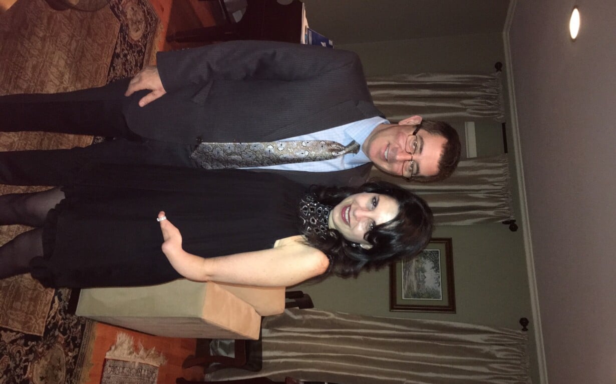 A man in a suit and tie stands next to a woman in a black dress with a lace neckline. They are smiling, posing in a warmly lit room adorned with patterned rugs and curtains, reminiscent of settings used for diversity programs aimed at fostering understanding through shared experiences.
