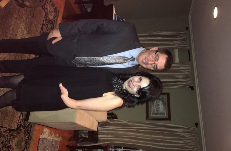 A man in a suit and tie stands next to a woman in a black dress with a lace neckline. They are smiling, posing in a warmly lit room adorned with patterned rugs and curtains, reminiscent of settings used for diversity programs aimed at fostering understanding through shared experiences.