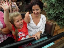 A woman and a young girl, embodying the joy of social emotional learning activities, ride a roller coaster with smiles and raised hands. The background shows other passengers and greenery, creating a vibrant setting for this shared experience.