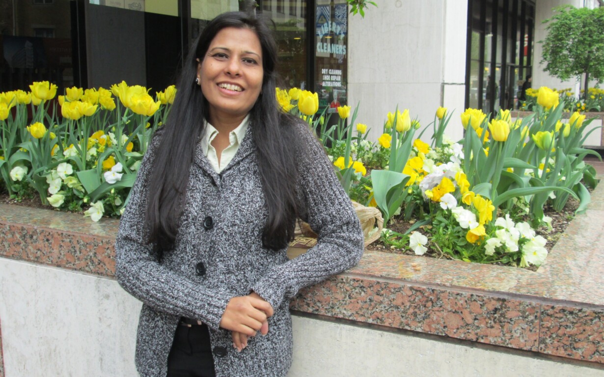 A person is smiling and leaning against a planter filled with yellow tulips and white flowers, embodying the warmth of social emotional learning activities. They are wearing a gray sweater in an urban setting with buildings and trees as a backdrop.