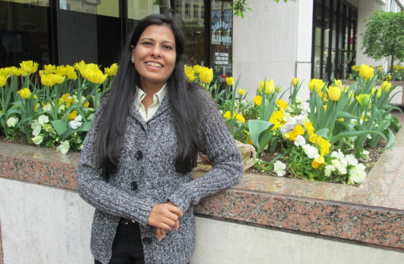 A person is smiling and leaning against a planter filled with yellow tulips and white flowers, embodying the warmth of social emotional learning activities. They are wearing a gray sweater in an urban setting with buildings and trees as a backdrop.