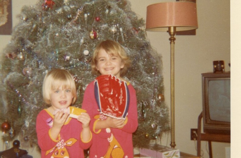 Two children in matching Christmas pajamas stand in front of a decorated Christmas tree, embodying the spirit of togetherness akin to social emotional learning activities. One holds a toy, the other a red baseball glove. Gifts are scattered around, with a vintage TV and lamp in the background.