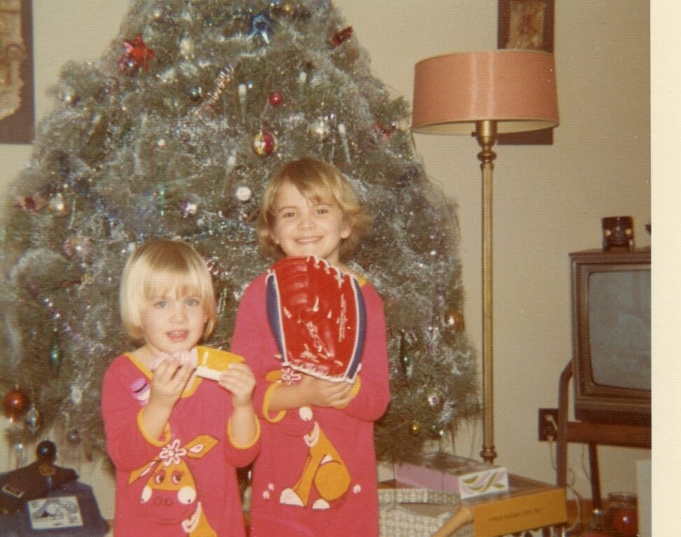 Two children in matching Christmas pajamas stand in front of a decorated Christmas tree, embodying the spirit of togetherness akin to social emotional learning activities. One holds a toy, the other a red baseball glove. Gifts are scattered around, with a vintage TV and lamp in the background.