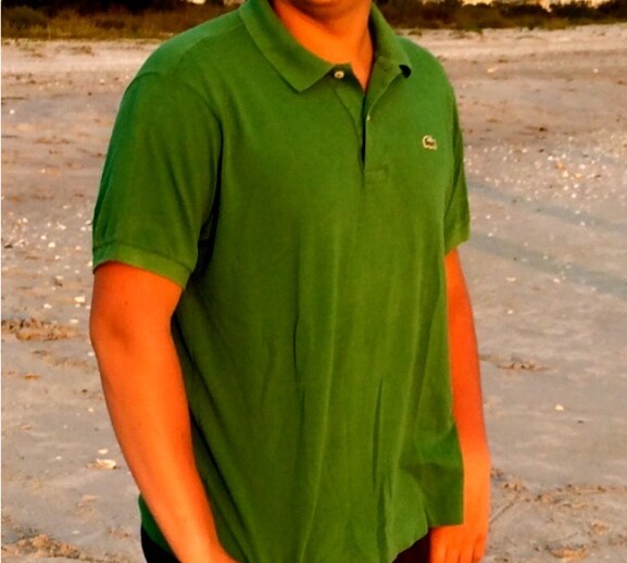 A person in a green shirt and dark shorts is standing on a sandy beach, smiling broadly. The background features distant houses and a clear blue sky, fostering the perfect setting for SEL activities that promote joy and connection.