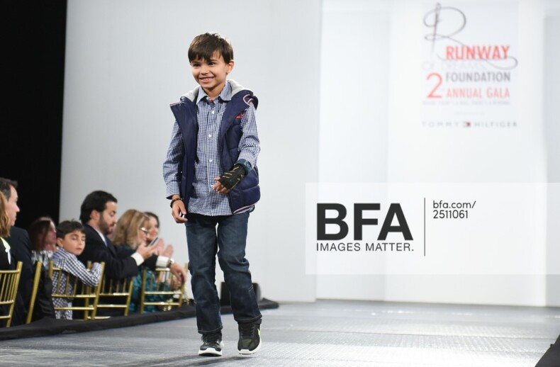 A young boy in a casual outfit confidently walks the runway, smiling. He's in a checkered shirt, jeans, and sneakers with a vest.  Part of a diversity program, the audience watches from the sides as an event banner is visible in the background.