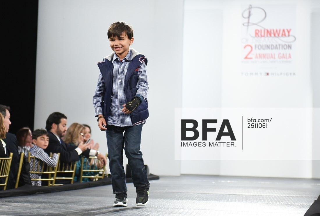 A young boy in a casual outfit confidently walks the runway, smiling. He's in a checkered shirt, jeans, and sneakers with a vest.  Part of a diversity program, the audience watches from the sides as an event banner is visible in the background.