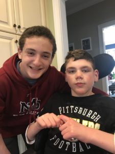 Two boys are smiling in a home setting, engaging in what could be a get-to-know-you activity. One is seated in a wheelchair wearing a black Pittsburgh shirt, while the other leans in, sporting a maroon hoodie. They seem to be in the kitchen area, cabinets visible behind them.