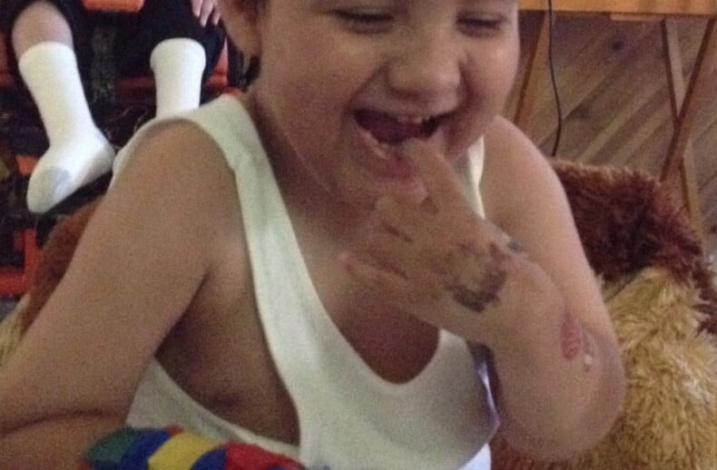 A young child with short hair smiles joyfully, sitting at a table engaged in SEL activities. Their arm is wrapped in a colorful bandage as they touch stickers on the table, part of get-to-know-you activities for students. Another person is seated in the background.