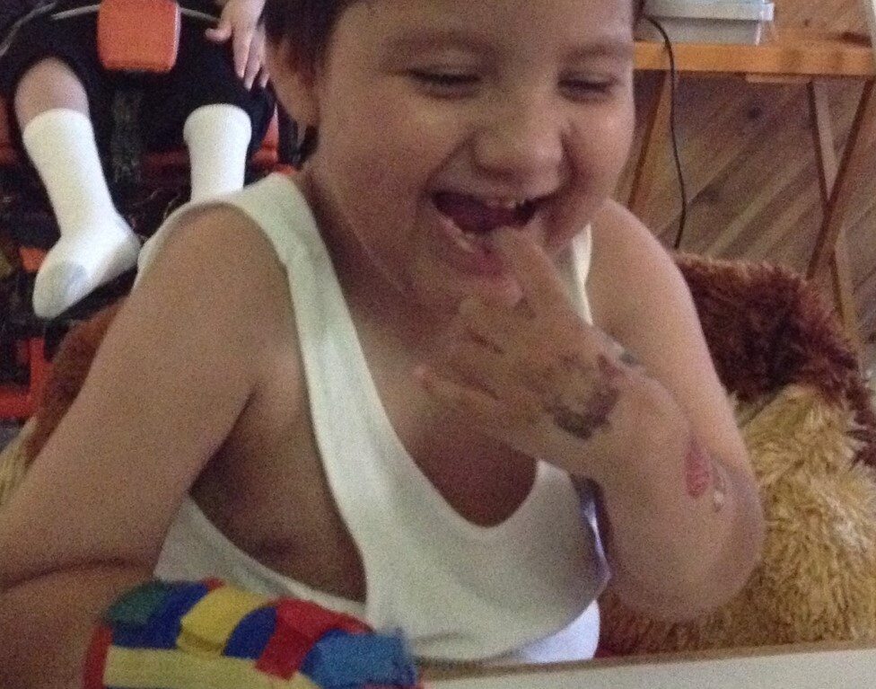A young child with short hair smiles joyfully, sitting at a table engaged in SEL activities. Their arm is wrapped in a colorful bandage as they touch stickers on the table, part of get-to-know-you activities for students. Another person is seated in the background.