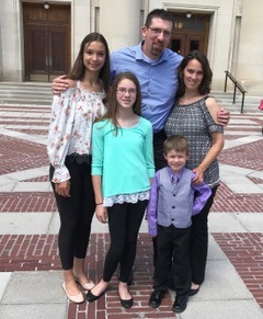 A family of five stands outdoors on a patterned brick walkway, embodying the spirit of diversity programs. The group includes an adult man and woman, two girls, and a young boy. They smile warmly in their casual and semi-formal attire, with a building entrance providing a picturesque backdrop.