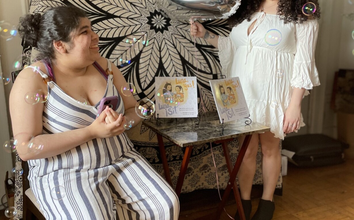 Two women celebrating indoors with bubbles, holding a large silver number "2" balloon. One wears a white dress and the other a striped jumpsuit. Amidst the festivities, their joy resembles that of students engaging in get-to-know-you activities. A decorative mandala and framed illustrations adorn the background.