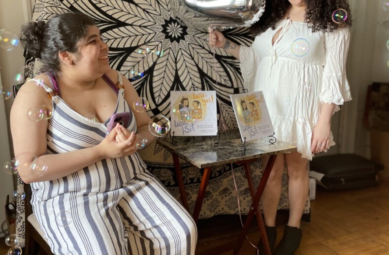 Two women celebrating indoors with bubbles, holding a large silver number "2" balloon. One wears a white dress and the other a striped jumpsuit. Amidst the festivities, their joy resembles that of students engaging in get-to-know-you activities. A decorative mandala and framed illustrations adorn the background.
